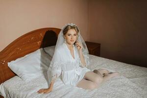 ein erwachsen Braut sitzt auf das Bett im ihr Zimmer mit nackt Beine im ein Weiß Kleid. Herrlich Hochzeit Haar und bilden foto