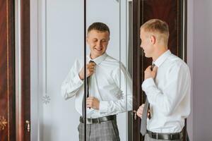 das Bräutigam setzt auf seine Krawatte im seine Zimmer und bereitet vor zum das Hochzeit. das Mann trägt ein Krawatte foto