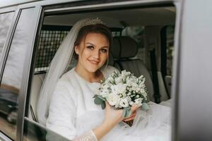 ein schön lächelnd Braut auf ihr Hochzeit Tag ist Sitzung im ein Auto und halten ein Hochzeit Strauß von Weiß und Rosa Rosen. ein jung Frau mit ein schön Frisur freut sich auf ihr Hochzeit Tag foto