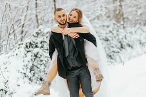 schön Hochzeit Paar Gehen im Winter schneebedeckt Wald, Frau im Weiß Kleid und Nerz Pelz Mantel, bärtig Mann im schwarz Mantel. das Bräutigam trägt das Braut im seine Waffen, ein komisch Foto