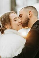 Hochzeit im Winter. stilvoll Braut im Weiß Nerz Mantel und Bräutigam im schwarz Mantel im Wald mit Weiß Schnee im kalt Wetter. das Bräutigam Küsse das Braut gegen ein verschwommen Hintergrund foto