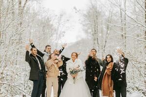 glücklich Jungvermählten und ihr freunde im Winter Mäntel Stand im ein schneebedeckt Wald und werfen Schnee mit ihr Hände foto