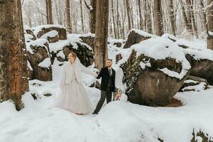 Hochzeit im Winter. stilvoll Braut im ein Nerz Mantel und Bräutigam im ein Mantel, im ein Wald mit Weiß Schnee im kalt Wetter. das Braut und Bräutigam sind Gehen unter Felsen und groß Steine im das Wald foto