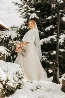 ein lächelnd und glücklich Braut auf das Hintergrund von schneebedeckt Weihnachten Bäume mit ein üppig Strauß von Blumen im ihr Hände. Porträt von ein schön Braut im ein Weiß Poncho und Hochzeit Kleid im ein Winter Park foto