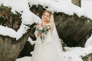 glücklich Braut auf das Hintergrund von schneebedeckt Felsen mit ein üppig Strauß von Blumen im ihr Hände und ein verschwommen Hintergrund. Porträt von ein schön Braut im ein Poncho im das kalt Jahreszeit foto