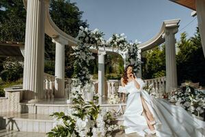 schön Braut auf Hintergrund von Blumen, attraktiv Frau im modisch Hochzeit Kleid Sitzung draußen. glücklich Braut Frau. Braut mit lockig lange Haar. Hochzeit Tag. Hochzeit. foto