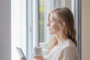 Frau Stehen neben das Fenster mit Handy, Mobiltelefon Telefon halten Kaffee Tasse foto