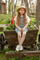 traurig wenig Mädchen Sitzung auf alt hölzern Wagen benutzt wie Blume Bett im Sommer- Park foto