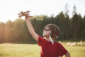 glückliches Kind im Pilotenhelm, das mit einem hölzernen Spielzeugflugzeug spielt und davon träumt, fliegen zu werden