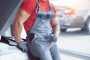 Der hübsche junge Automechaniker in Uniform untersucht das Auto im Autoservice foto