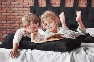 zwei Kinder liegen auf einem großen Bett und lesen ein interessantes Buch. sie tragen den gleichen Pyjama