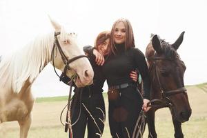 zwei junge schöne mädchen in ausrüstung zum reiten in der nähe ihrer pferde. sie lieben tiere