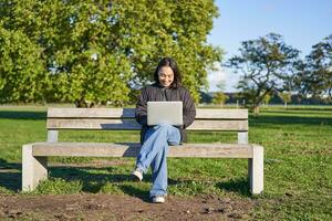 Porträt von jung asiatisch Frau Arbeiten auf Laptop, Sitzung auf Bank im Park auf sonnig Tag, mit ihr Computer, studieren im ziemlich Platz foto