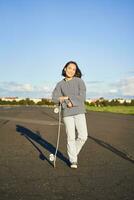 Vertikale Porträt von lächelnd asiatisch Frau Stehen auf Straße mit Longboard, Skateboarding auf lange Kreuzer, posieren auf leeren Straße auf sonnig Tag foto