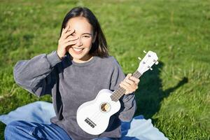 lächelnd asiatisch Mädchen mit Ukulele, spielen im Park und Singen, Lebensstil Konzept foto