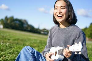 glücklich Menschen und Hobbys. lächelnd asiatisch Mädchen spielen Ukulele Gitarre und Singen, Sitzung im Park draußen auf Decke foto