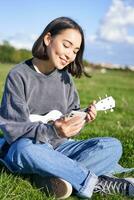 Vertikale Schuss von lächelnd asiatisch Mädchen mit Smartphone, spielen Ukulele, lesen Akkorde oder Text während Singen, entspannend draußen. Lebensstil und Menschen Konzept foto