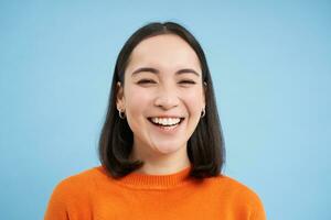 schließen oben Porträt von schön Lachen asiatisch Frau, zeigt an offen Glück, lächelnd beim Kamera, Stehen Über Blau Hintergrund foto