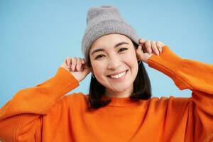 schließen oben Porträt von lächelnd asiatisch Frau im warm Hut, suchen glücklich und süß beim Kamera, hat klar natürlich Haut, steht Über Blau Hintergrund foto