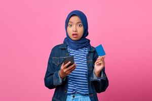 Porträt einer schockierten jungen Asiatin, die Smartphone hält, während sie Kreditkarte zeigt