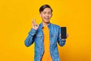 Porträt eines jungen asiatischen Mannes, der Smartphone hält und ein OK-Zeichen auf gelbem Hintergrund zeigt