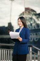 zuversichtlich asiatisch Frau mit ein Lächeln Stehen halten Notizblock und Tablette beim aus Seite Büro. groß Stadt foto