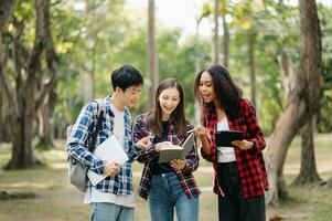 drei jung Hochschule Studenten ist lesen ein Buch während entspannend Sitzung auf Gras im ein Campus Park mit ihr Freunde. Bildung Konzept foto