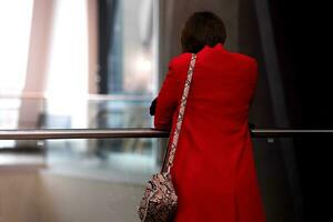 jung Frau im ein rot Mantel steht mit ihr zurück zu das Zuschauer im das Empfangshalle von ein Öffentlichkeit Gebäude foto