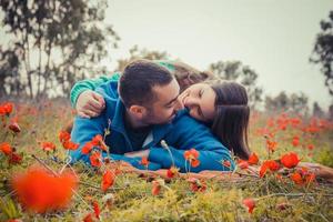 junges Paar liegt auf dem Gras in einem Feld aus roten Mohnblumen und lächelt sich an foto