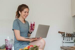 schön jung Frau im Übung Kleider haben Spaß im ein süß Küche beim heim. mit Ihre Laptop zu bereiten vegan Obst Salat Dressing, Obst zittert, oder gesund Smoothies. foto