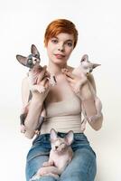 Rothaarige jung Frau halten zwei Sphynx Katze im Hände und einer Kätzchen Lügen auf ihr Beine. ziemlich Frau mit kurz Haar im T-Shirt und Jeans suchen Kamera, Lügen Nieder auf Weiß Hintergrund. Teil von Serie foto