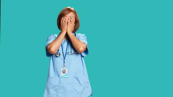 müde bipoc erfahren Krankenschwester Abdeckung Gesicht mit Palmen, erschöpft nach lange Tag beim arbeiten. asiatisch Gesundheitswesen Fachmann im brauchen von brechen von Arbeit, isoliert Über Blau Studio Hintergrund foto