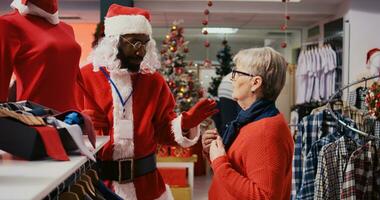 Mitarbeiter tragen Santa claus Kostüm im beschäftigt Einkaufen Einkaufszentrum Mode Boutique chatten mit älter Frau. Arbeiter im Urlaub thematisch passen zeigen alt Kunde rot Kleidung Stück foto