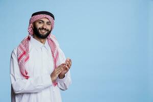 heiter lächelnd Muslim Person tragen traditionell Kleider klatschen Hände und suchen beim Kamera. aufgeregt arabisch Mann zeigen Beifall Geste Studio Porträt auf Blau Hintergrund foto