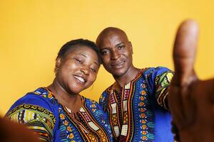 lächelnd schwarz Mann und Frau tragen ethnisch Kleider nehmen Selfie auf Smartphone und suchen beim Vorderseite Kamera. heiter Paar halten Handy, Mobiltelefon Telefon und Herstellung Foto pov Porträt