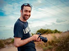 glücklicher junger kaukasischer Mannfotograf, der eine Digitalkamera in der Hand hält und in die Kamera im Freien im Feld lächelt