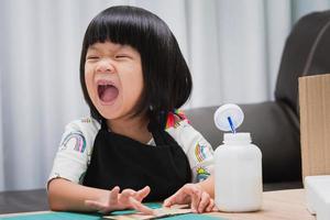 glückliches asiatisches Mädchen mit schwarzer Schürze, Kinderlachen beim Aufkleben auf ein Stück Handwerk. das Konzept des spielerischen Lernens. Kind im Alter von 4-5 Jahren. foto