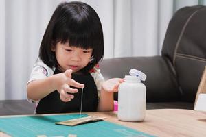 Erfindermädchen ist begeistert, dehnbaren Kleber auf Kartonstücke aus Karton aufzutragen. Kind viel Spaß mit kreativen Design-Werkstücken. Kindergarten mit schwarzer Schürze studiert im Klassenzimmer. foto