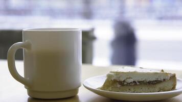 Nahansicht von Weiß Becher und Teller mit Käsekuchen. Morgen Frühstück mit Kaffee und klassisch Käsekuchen auf hölzern Tisch. Süss Frühstück im Cafe auf Hintergrund von Fenster mit beschäftigt Straße foto