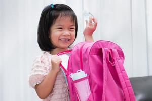 Ein süßes Mädchen hat Spaß beim Packen ihrer Taschen zur Schule. Mädchen, das eine Flasche Alkoholgel hält. die Hygienemaske befindet sich neben der Tasche.