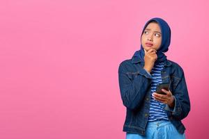 Porträt einer jungen asiatischen Frau, die mit der Hand am Kinn über eine Frage nachdenkt, während sie das Smartphone hält