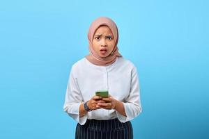 Porträt einer schockierten jungen Asiatin mit offenem Mund mit Handy über blauem Hintergrund foto