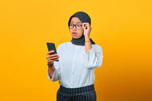 überraschte junge asiatische frau, die mit offenem mund auf gelbem hintergrund auf den smartphonebildschirm schaut