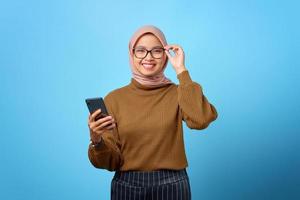 glückliche junge asiatische frau, die brille berührt und handy auf blauem hintergrund hält foto