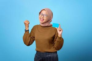 glückliche junge asiatische frau, die kreditkarte mit handgesteerfolg auf blauem hintergrund hält foto