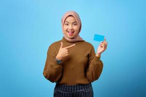 fröhliche junge asiatische frau, die mit dem finger auf die kreditkarte auf blauem hintergrund zeigt foto