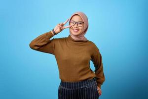 fröhliche junge asiatische frau, die friedenszeichen über auge auf blauem hintergrund zeigt foto