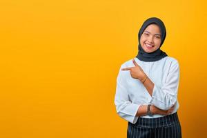 lächelnde junge asiatische frau, die mit den fingern auf den kopierraum zeigt und die kamera auf gelbem hintergrund schaut foto