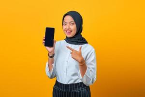 Porträt einer fröhlichen jungen asiatischen Frau, die einen leeren Bildschirm des Smartphones auf gelbem Hintergrund zeigt