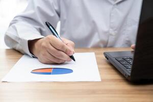 Mann verwenden Stift schreiben auf ein Graph Papier im Arbeit Prozess täglich auf Tabelle und verwenden Laptop Computer im Hintergrund foto